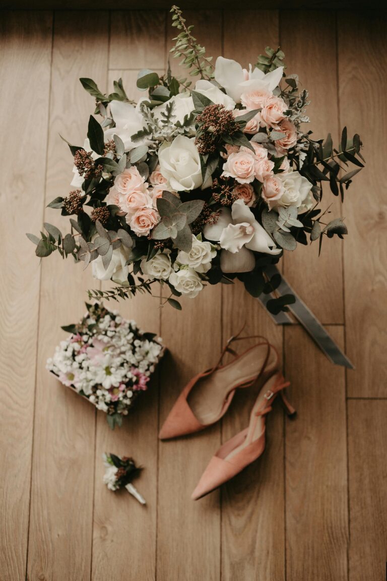 Top view of a bridal bouquet with wedding shoes and floral accessories on wood floor.